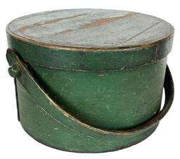 **SOLD** K1765 Early 19th century New England wooden bail handle Pantry Box bearing the original green painted surface. Heavy construction with tacked lap joints and a hand hewn steamed and bentwood swing handle that is secured with wooden pegs. The wooden pegs are secured inside the box with tiny wooden pins. Natural patina interior. Measurements: 11 ½ top diameter x 7 tall.