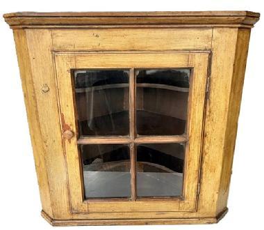 L161 Early 19th century Pennsylvania glass door hanging corner cupboard with applied molding on top and bottom bearing its original mustard painted surface. The four-windowpane door is fully mortised and wooden peg construction with a beaded edge and opens to reveal two butterfly shelves and one straight edge shelf inside. The wood is pine with square head nail construction. Cupboard is 29 ¼” tall and takes a 21 ½” deep corner.