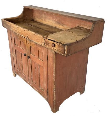 J127 Lancaster County, Pennsylvania original salmon painted drysink boasting rare Bible Cross Panel doors with beaded edges and a very unusual hinged shelf along the top that lifts to create a tall backsplash and accommodates larger items in the well area. One dovetailed drawer overhangs to the right-hand side of the drysink. Remnants of beaded edging remains along the drawer and apron of the dry sink. The notch cut out on the feet echoes the notched cut outs along the rounded sides of the well. Doors are triple mortised and wooden pegged. One board sides, and one board interior shelves, measure a full 20 wide/deep. Square head nail construction. Extremely dry surface with extensive wear indicating many years of heavy use. The wear on the shelf is indicative of use in both positions. Interior bears natural patina. Circa 1840s. Measurements: 53 ¼ wide x 22 ¼ deep x 42 tall with shelf down. It measures 46 ½ tall with shelf in the upright / backsplash position.