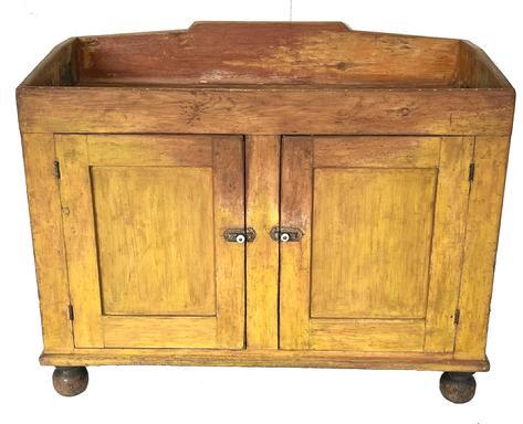**SOLD** J185 Early 19th century Lancaster County, Pennsylvania Drysink in original yellow painted surface featuring a deep well with tall shaped backsplash, two fully mortised and pegged doors, applied molding around the bottom and rests on applied turned feet bearing their original black painted surface. The bottom board of the drysink is fully dovetailed. This piece bears great wear from years of use. Faint remnants of a later salmon paint are evident in the areas of wear. The doors open to reveal a clean, natural patina interior with shelf for additional storage. Circa 1840s. Square head nail construction. Measurements: 44 wide x 20 ¾ deep x 36 tall (back) x 32 tall (front)