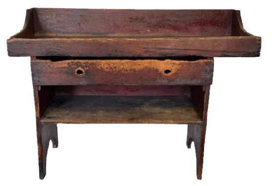 J217 Extremely rare Lancaster County, Pennsylvania bucket bench / dry sink featuring an overhanging well with a drawer and a mortised open shelf below the center section of the well. The surface retains traces of old gray paint over the original red paint, and bears extensive wear indicative of many years of repetitive use. The notched groove drain hole prominently remains in the overhang of the well. The drawer opens to reveal a divided section for utensils. One board, square head nail construction with nice, half-moon cut out ends. This is one of the earliest, and best, examples of the Lancaster County bucket bench / dry sinks we have seen in 40+ years. Circa 1830s 1840s. Measurements: 48 ¾ wide x 19 ½ deep x 35 ¼ tall.