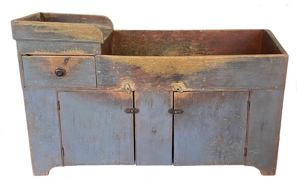 F146 Exceptional Lancaster County, Pennsylvania early 19th Century oversized Dry Sink in the original pewter gray paint with a bittersweet red painted well. Extremely unusual form having a 10 ½” deep well and slanted drain board top with incised grooves for water drainage. The drain board is wrapped with an applied dovetailed gallery. There is one oversized, dovetailed drawer over two plank doors. Each door has a batton on the inside to keep them from warping. The left side interior has one shelf, the right side interior is open for water bucket storage. Simple cut out foot. Measurements: 61” wide x 38” tall x 21 ¼” deep.